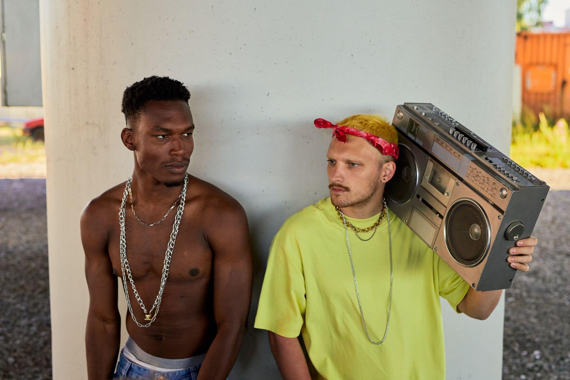 Two young men in casual attire with chains and a boombox outdoors.